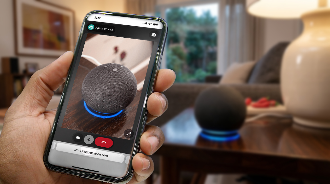 A customer holds up a phone in a live video session with an agent, its camera pointed at a smart speaker on a side table